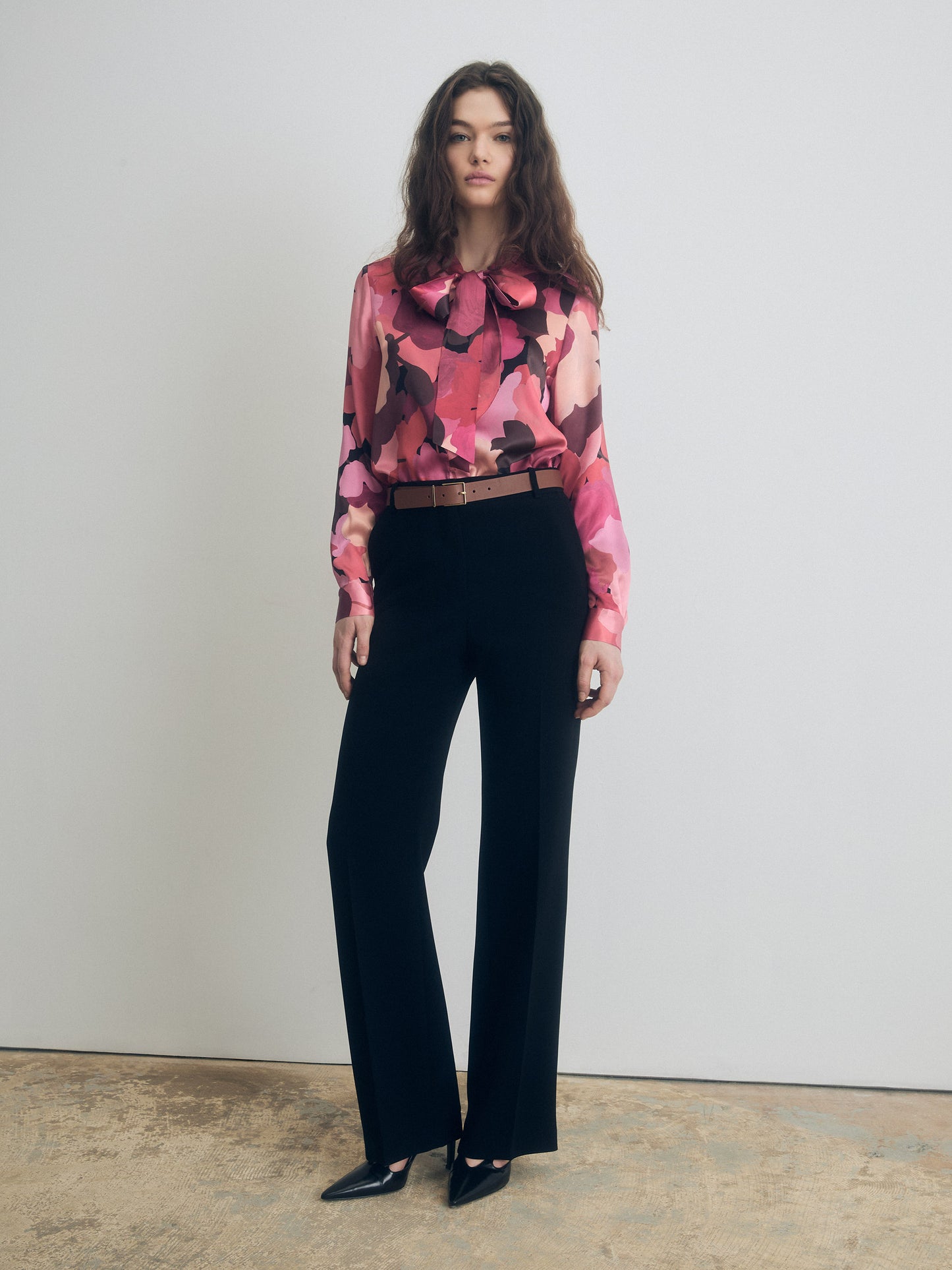 Front view of a long-sleeve silk blouse with abstract pink and burgundy print and a bow tie neckline, styled with high-waisted black tailored trousers and pointed-toe heels.