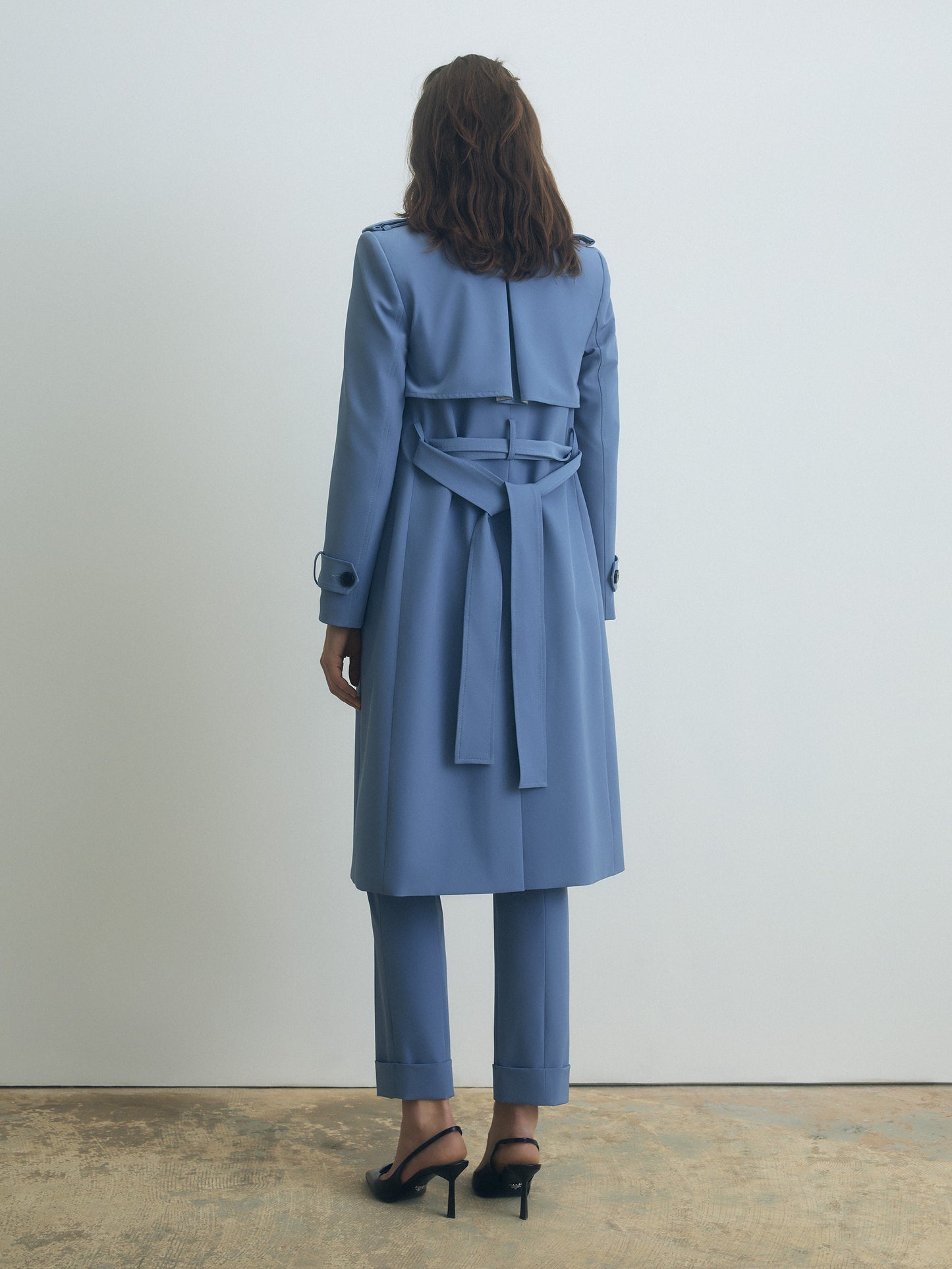Person wearing a light blue trench coat and pants standing against a white wall.