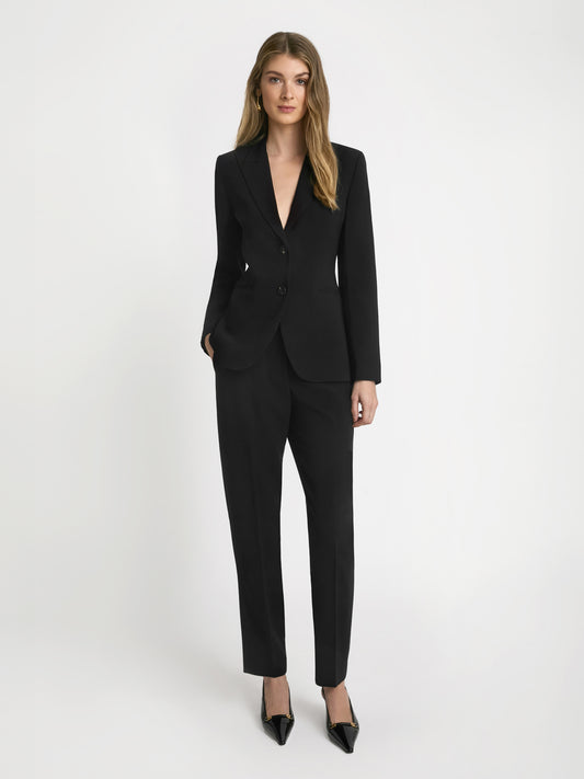 A woman standing in a studio setting, wearing a black slim pant suit with a blazer and heels.