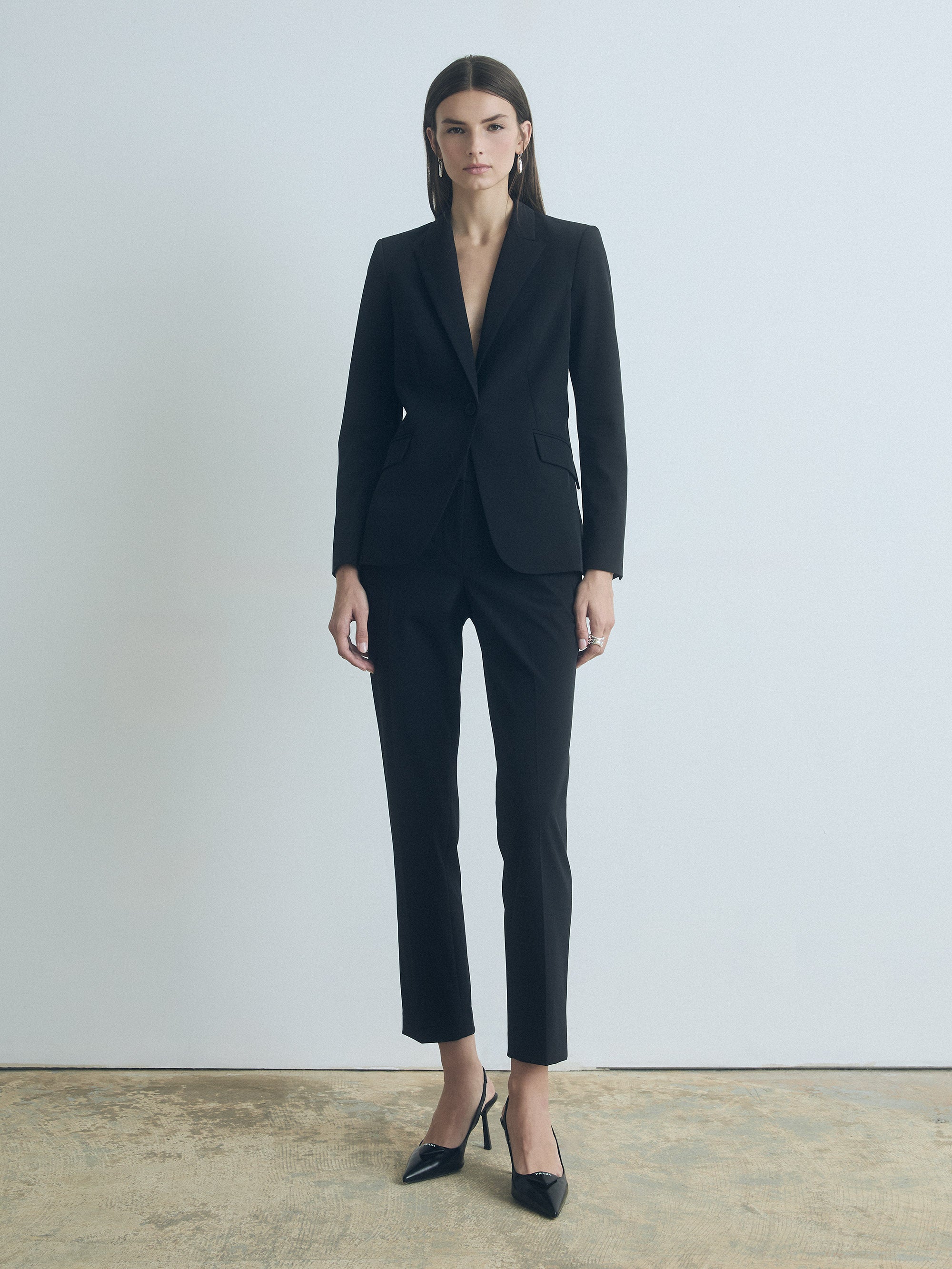 A model wearing black ankle length slim black pants with mid-rise waist, featuring slash pockets, back welt pockets, hook and eye closure with zip, and belt loops.