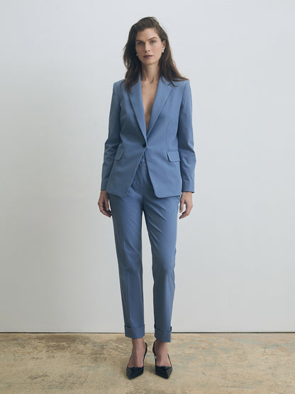 Model wearing a tailored smokey‑blue pantsuit featuring a fitted single‑button blazer with notched lapels and flap pockets, paired with slim cuffed trousers and black pointed‑toe heels. The look is photographed against a minimal light grey background
