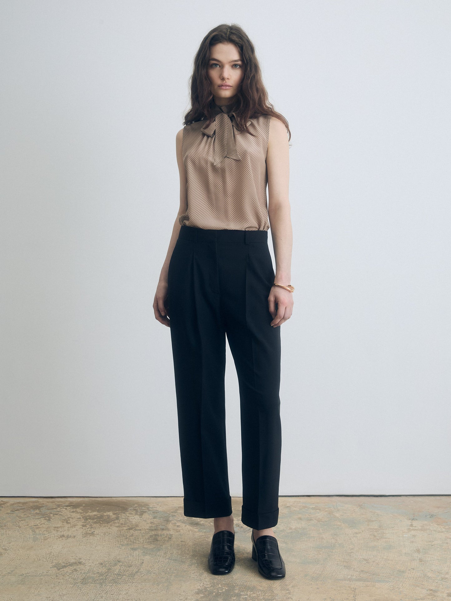  Woman wearing a black pleated cuffed trousers with a clean tailored waistband, slash pockets, and back welt pockets with a sleeveless blouse