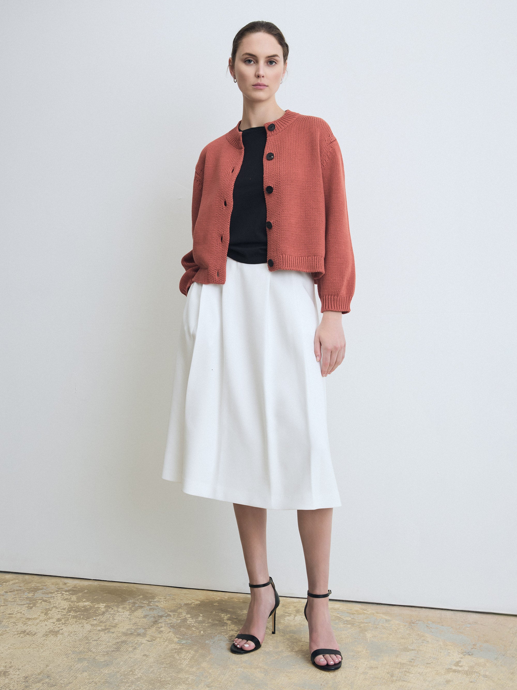 Woman wearing a terracotta cardigan, black top, white skirt, and black sandals on a plain background