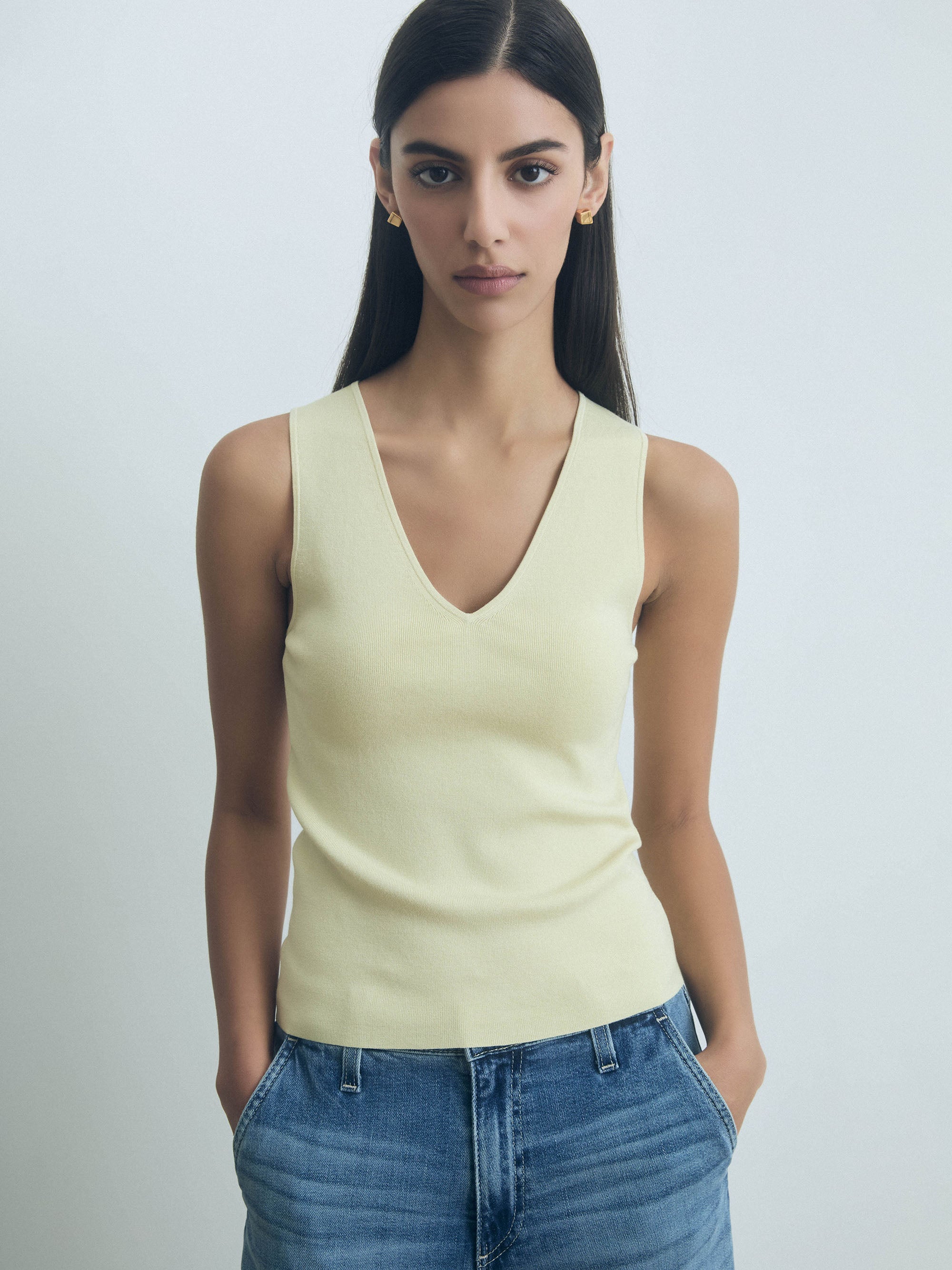 Woman wearing a light yellow sleeveless top and blue jeans against a plain background