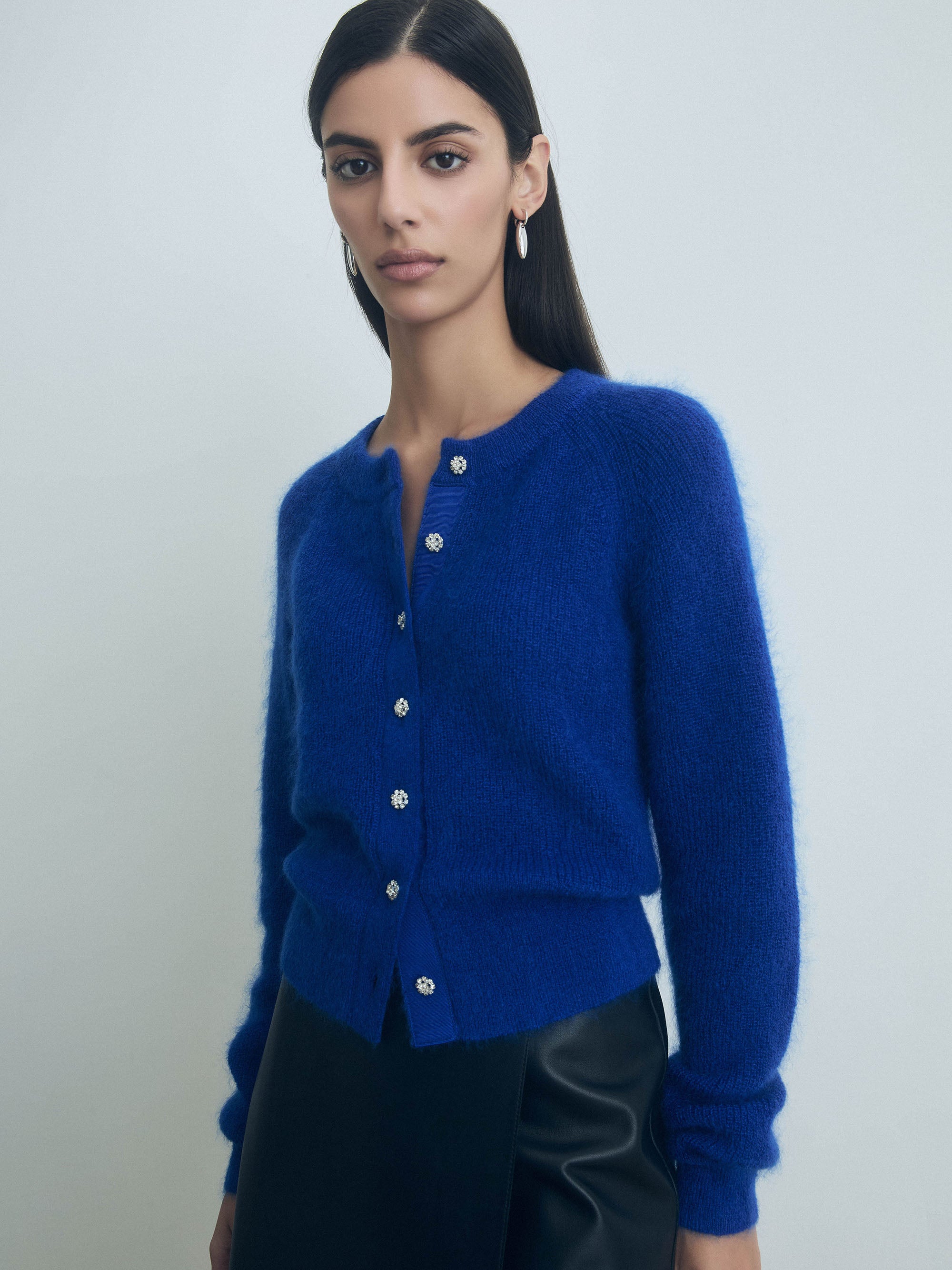 Woman wearing a blue judith and charles cardigan with embellished front button closure against a plain background