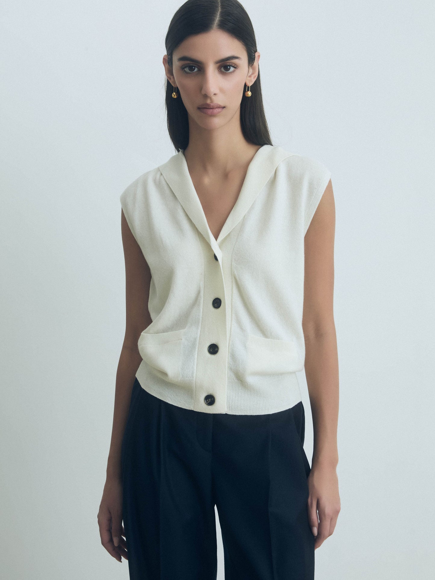 Woman wearing a Off White sleeveless hooded wool-cashmere vest with button front closure by Judith & Charles with black pants on a plain background