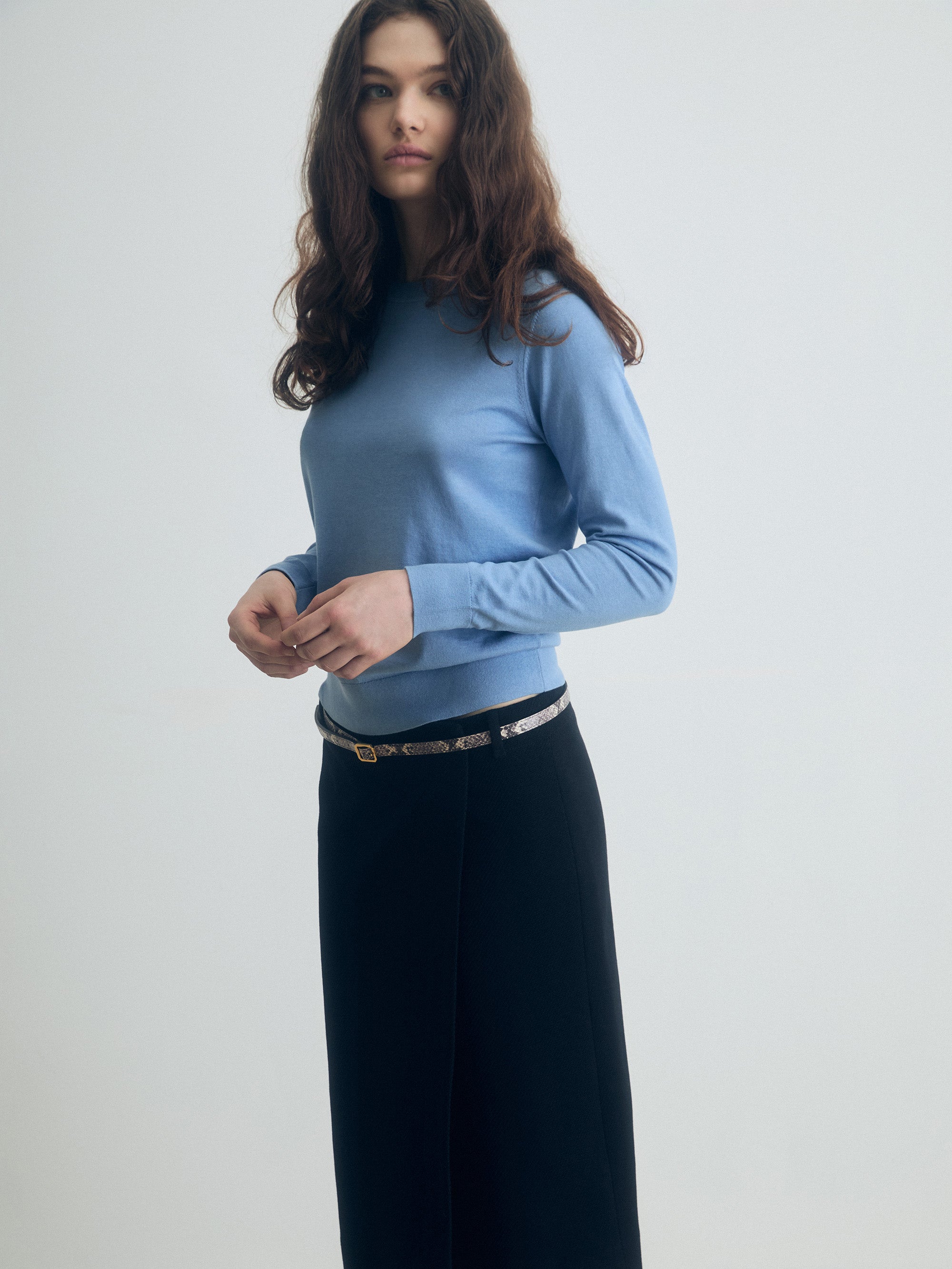 Smokey blue silk‑cotton crewneck sweater with long sleeves, styled with a black skirt and slim belt, shown on model.