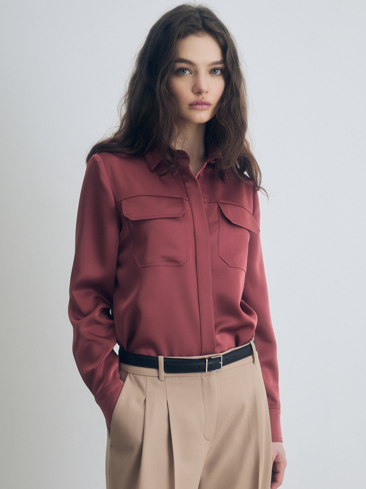 Terracotta silk blouse with flap chest pockets, button-down front, and long sleeves, styled with high-waisted beige tailored trousers and a black leather belt.