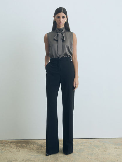 Woman wearing a sleeveless judith and charles top with a bow and dark pants against a plain background