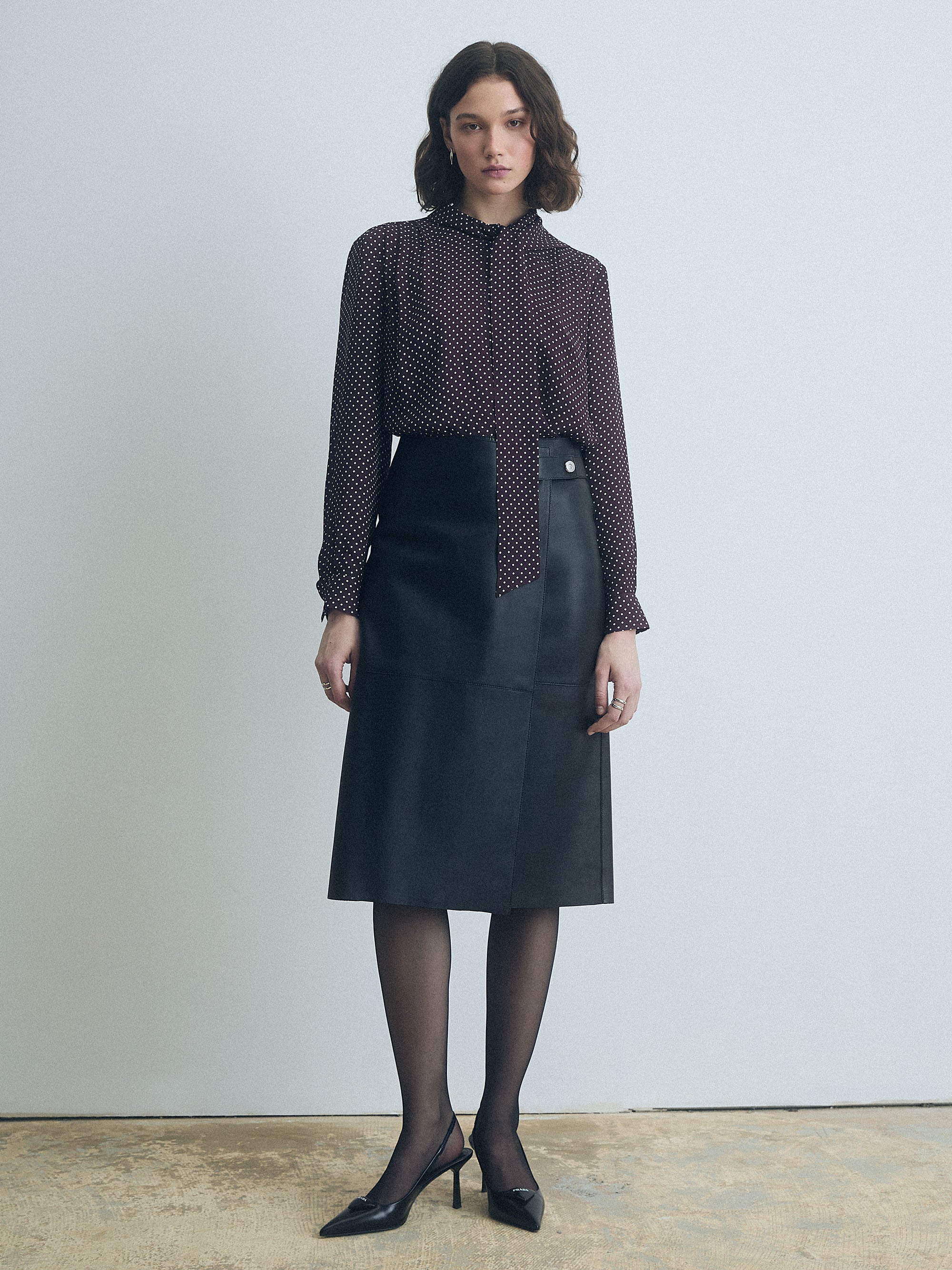 Woman wearing a dark cherry polka dot blouse and black leather judith and charles skirt against a plain background