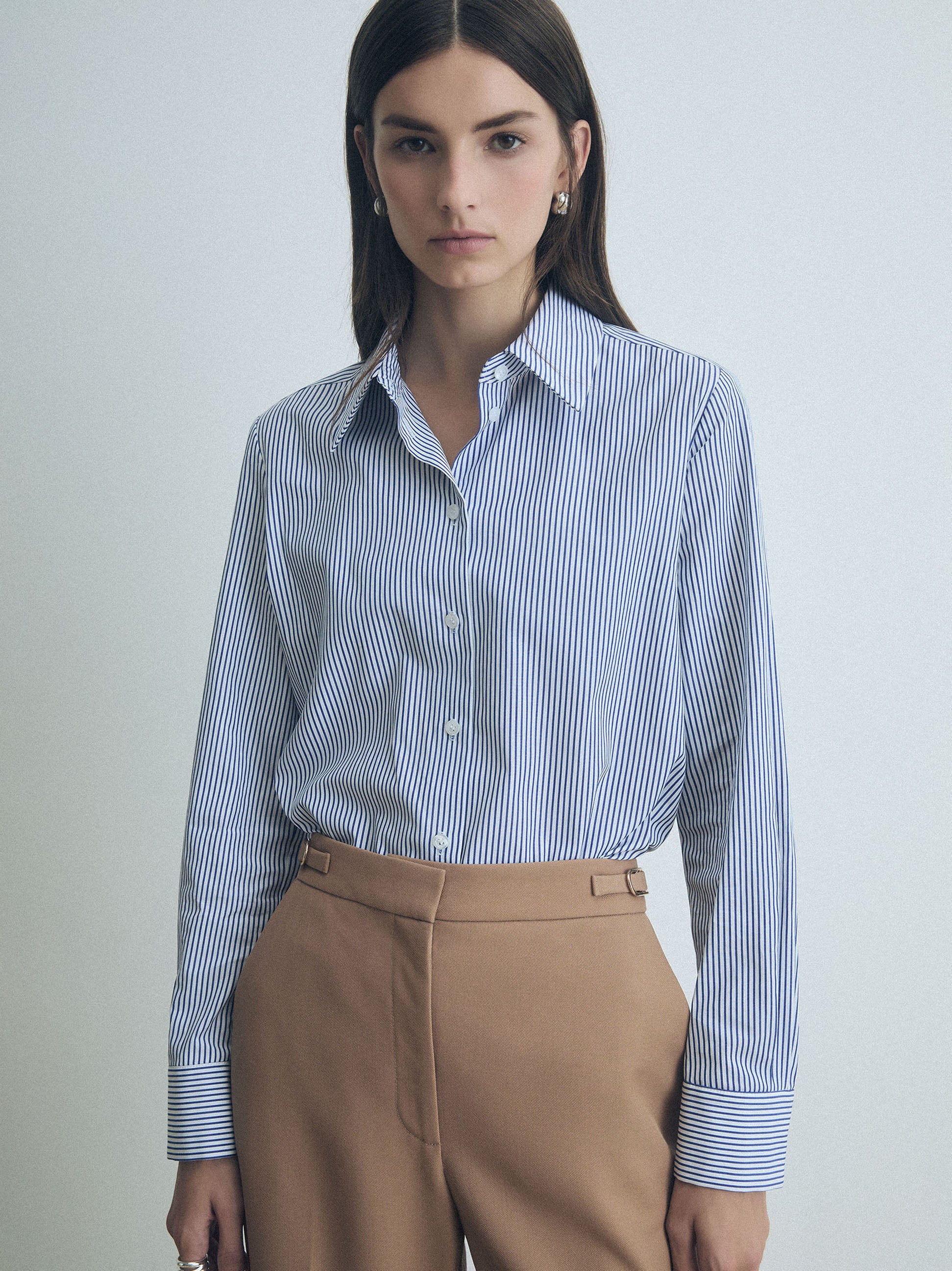 Woman wearing a blue and white striped judith and charles shirt with camel pants on a light gray background
