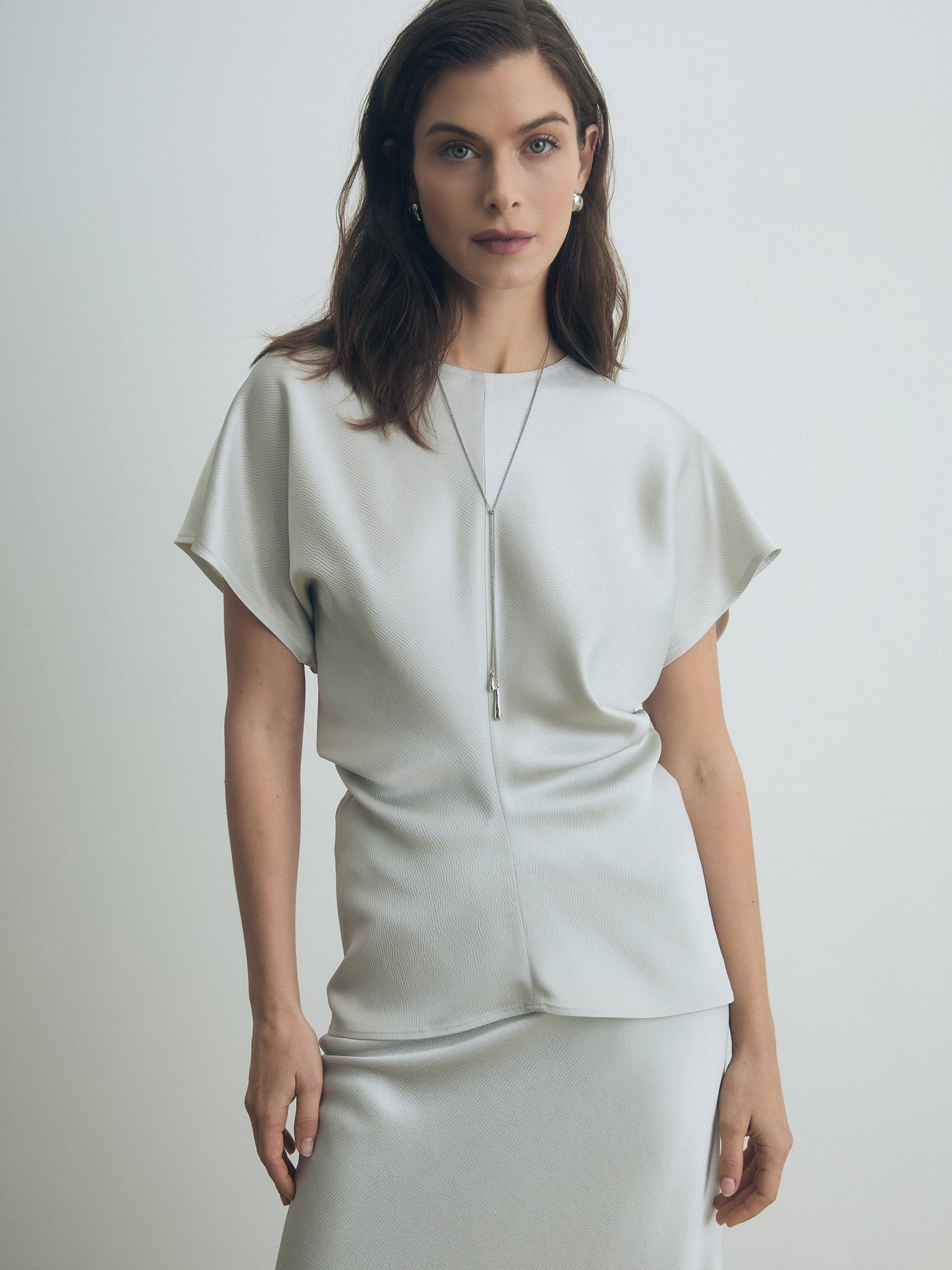 Model wearing a pearl satin cap sleeve blouse with crew neck and subtle side gathers, styled with a matching skirt. Minimalist, elegant silhouette.
