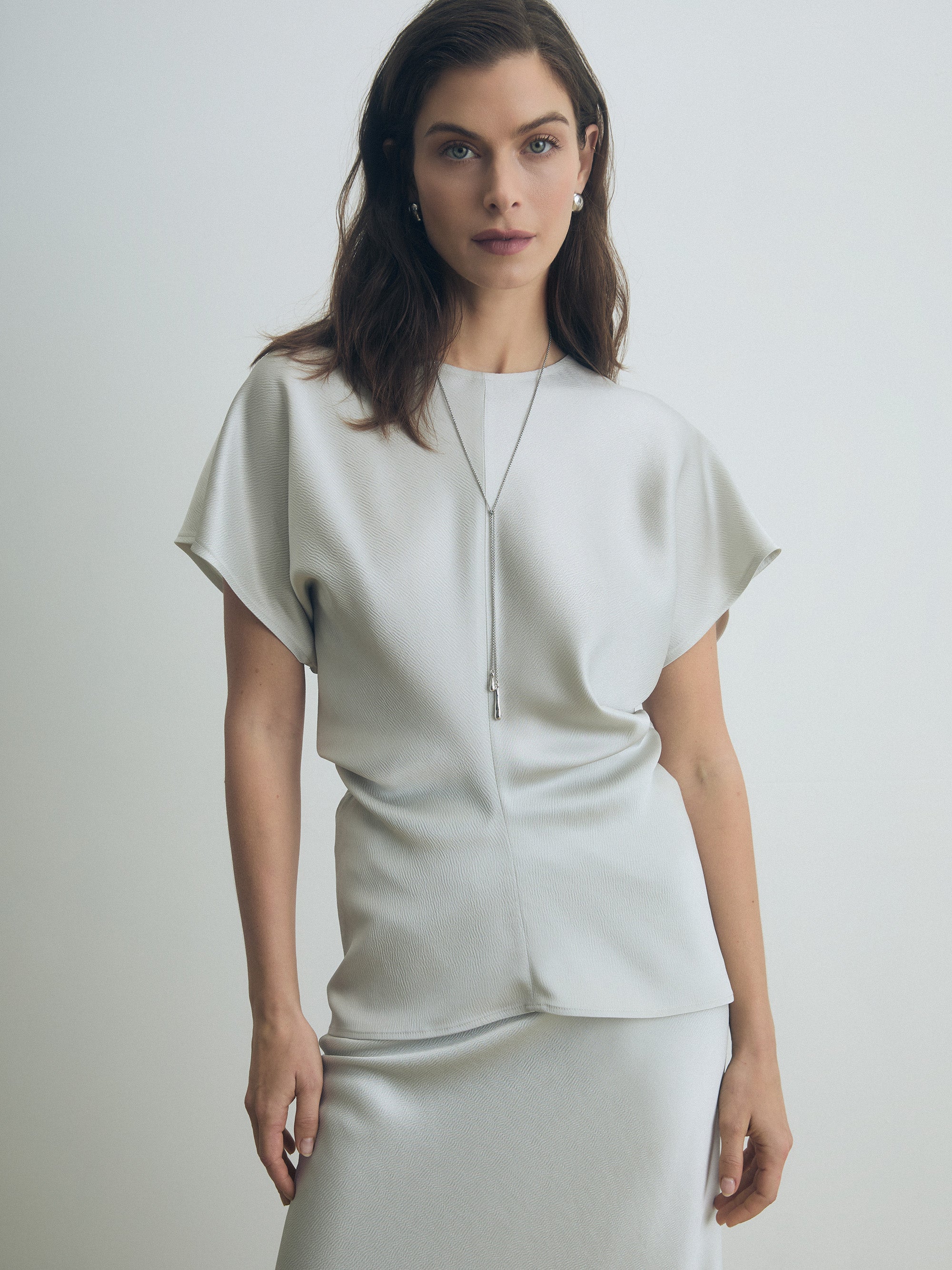 Model wearing a pearl satin cap sleeve blouse with crew neck and subtle side gathers, styled with a matching skirt. Minimalist, elegant silhouette.