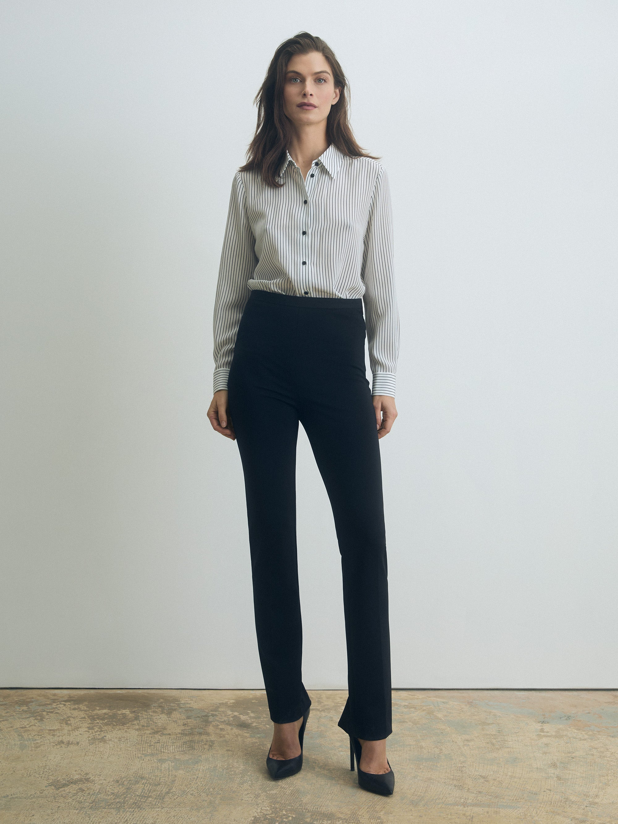 Model wearing a fitted long-sleeve blouse in black-and-white vertical stripes with a spread collar and front button closure, paired with high-waisted black tailored trousers and black pointed-toe heels