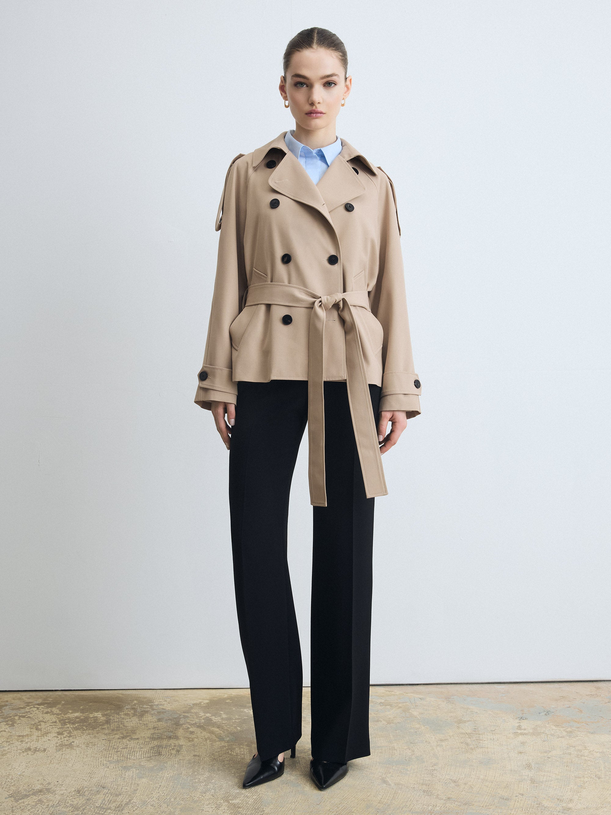 Woman wearing a beige trench coat over a blue shirt with black pants on a plain background