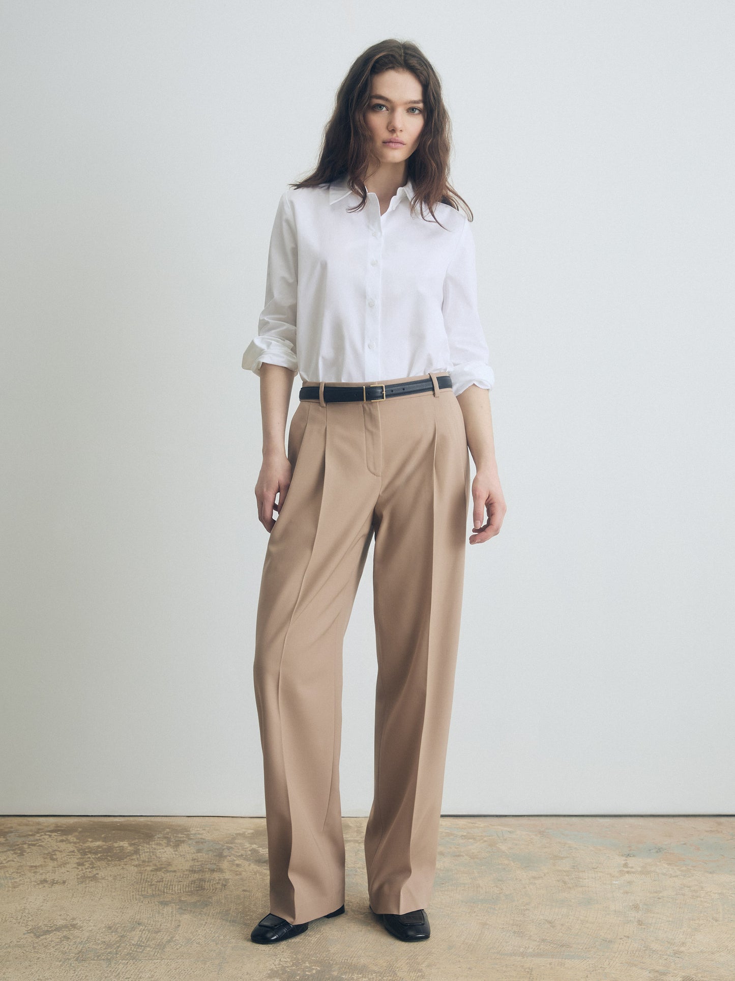 Woman wearing a white shirt and camel pants against a plain background