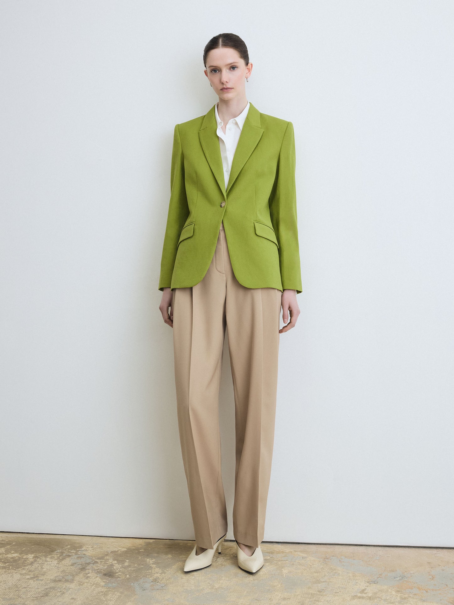 Person wearing a green blazer and beige pants against a white wall