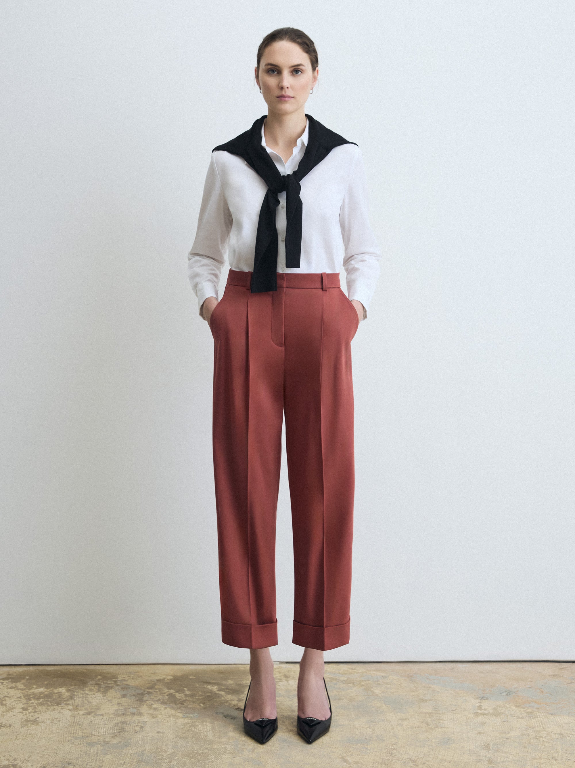 Woman wearing a white blouse with a black collar, terracotta pants, and black shoes on a plain background
