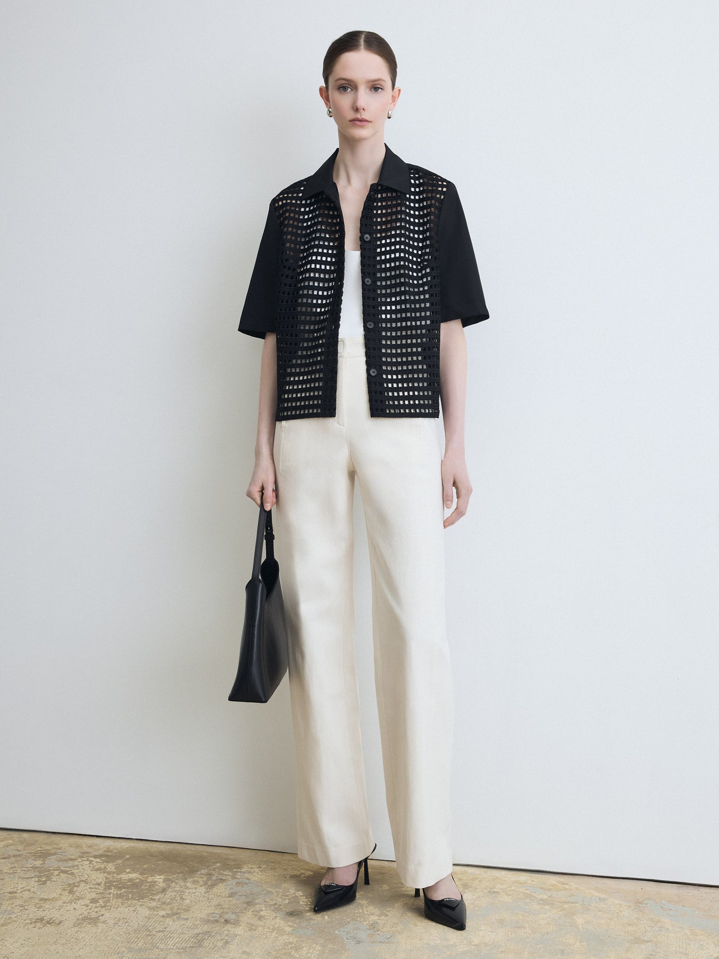 Woman wearing a black open-knit top and white pants, holding a black handbag against a plain background.