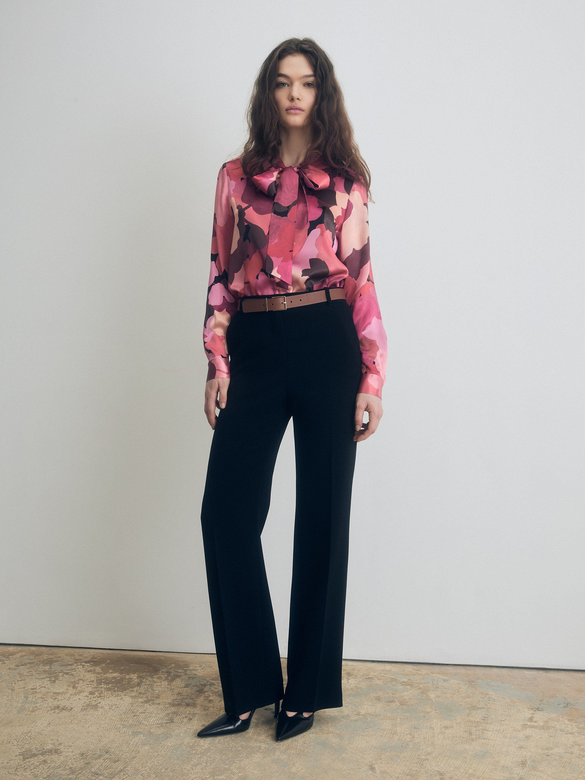 Front view of a long-sleeve silk blouse with abstract pink and burgundy print and a bow tie neckline, styled with high-waisted black tailored trousers and pointed-toe heels.