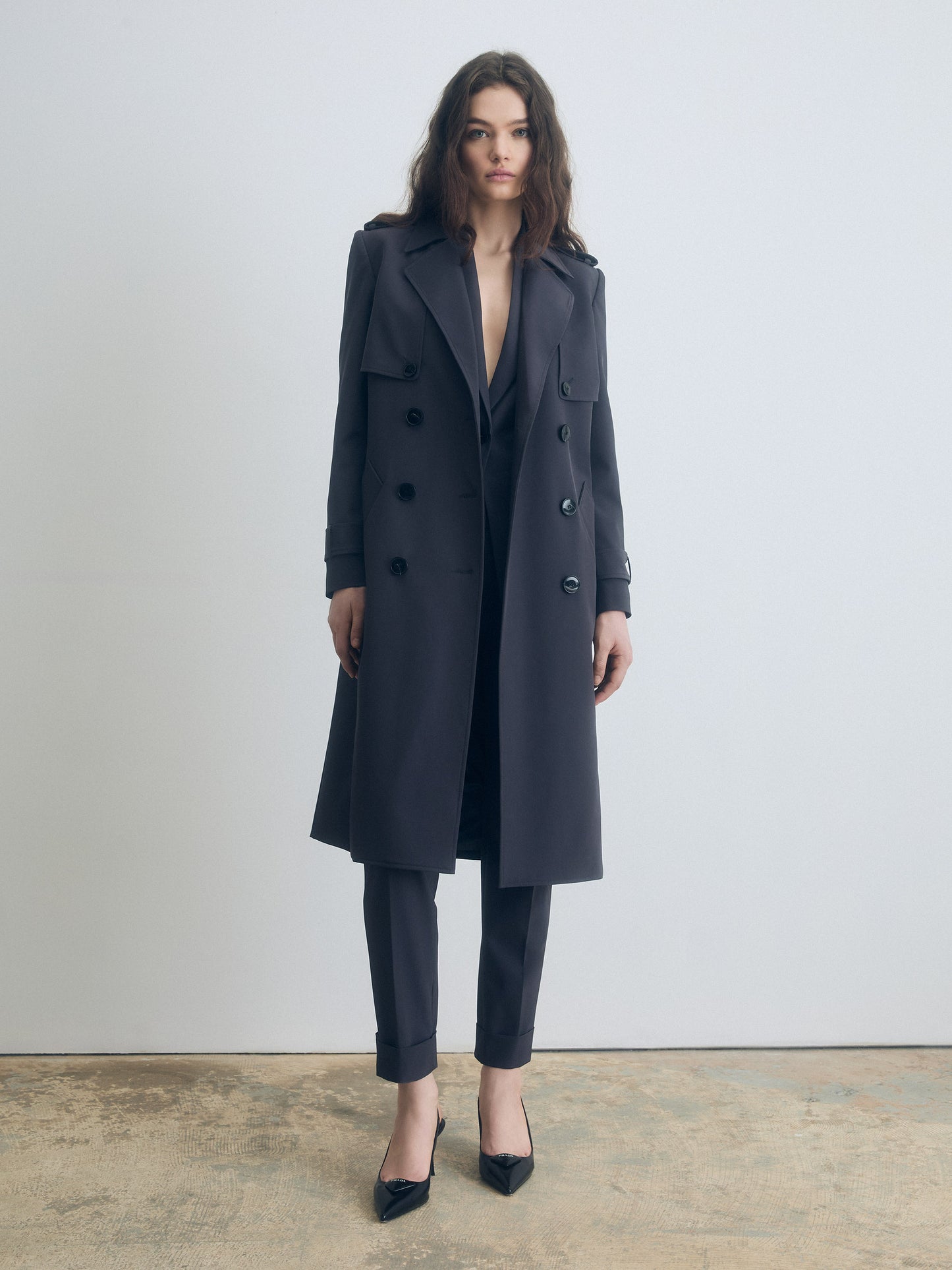 Woman wearing a long dark grey coat over a suit on a plain background