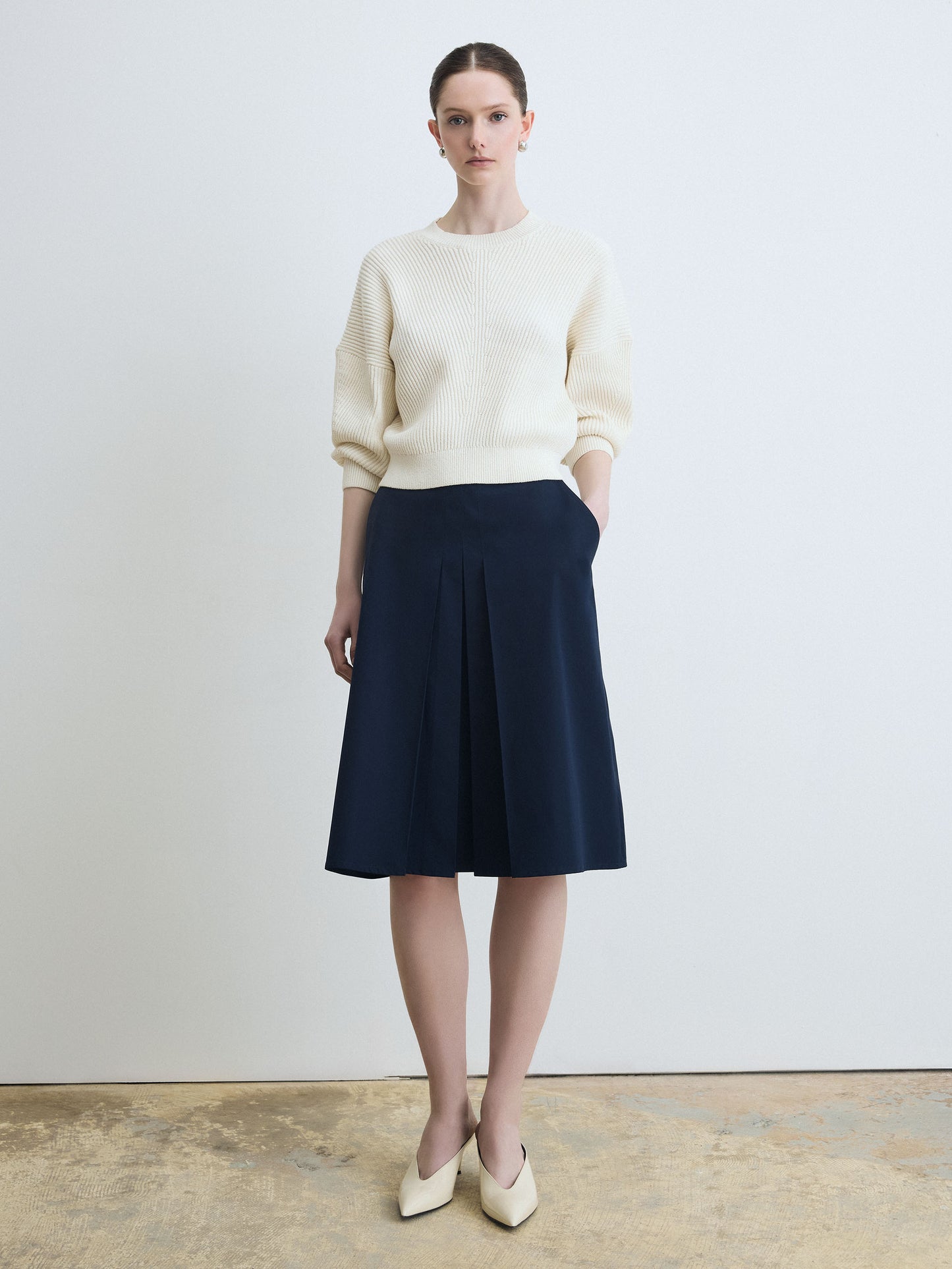 Woman wearing a cream sweater and navy skirt against a plain background