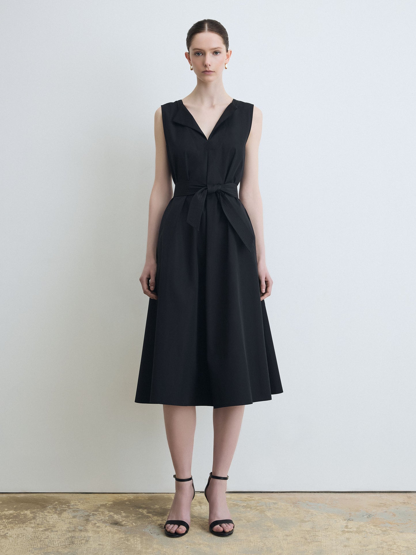 Woman wearing a black sleeveless dress with a belt against a plain background