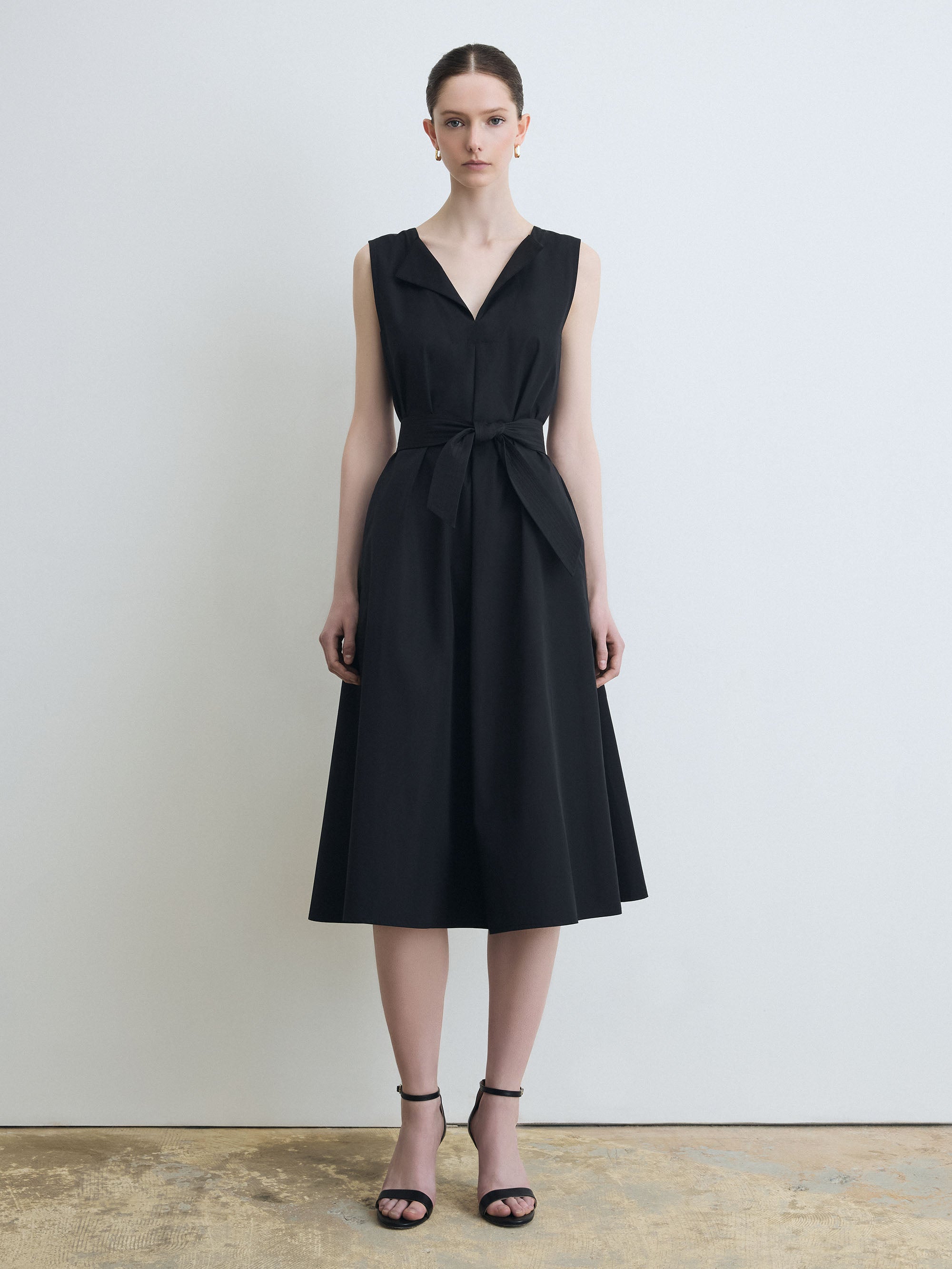 Woman wearing a black sleeveless dress with a belt against a plain background