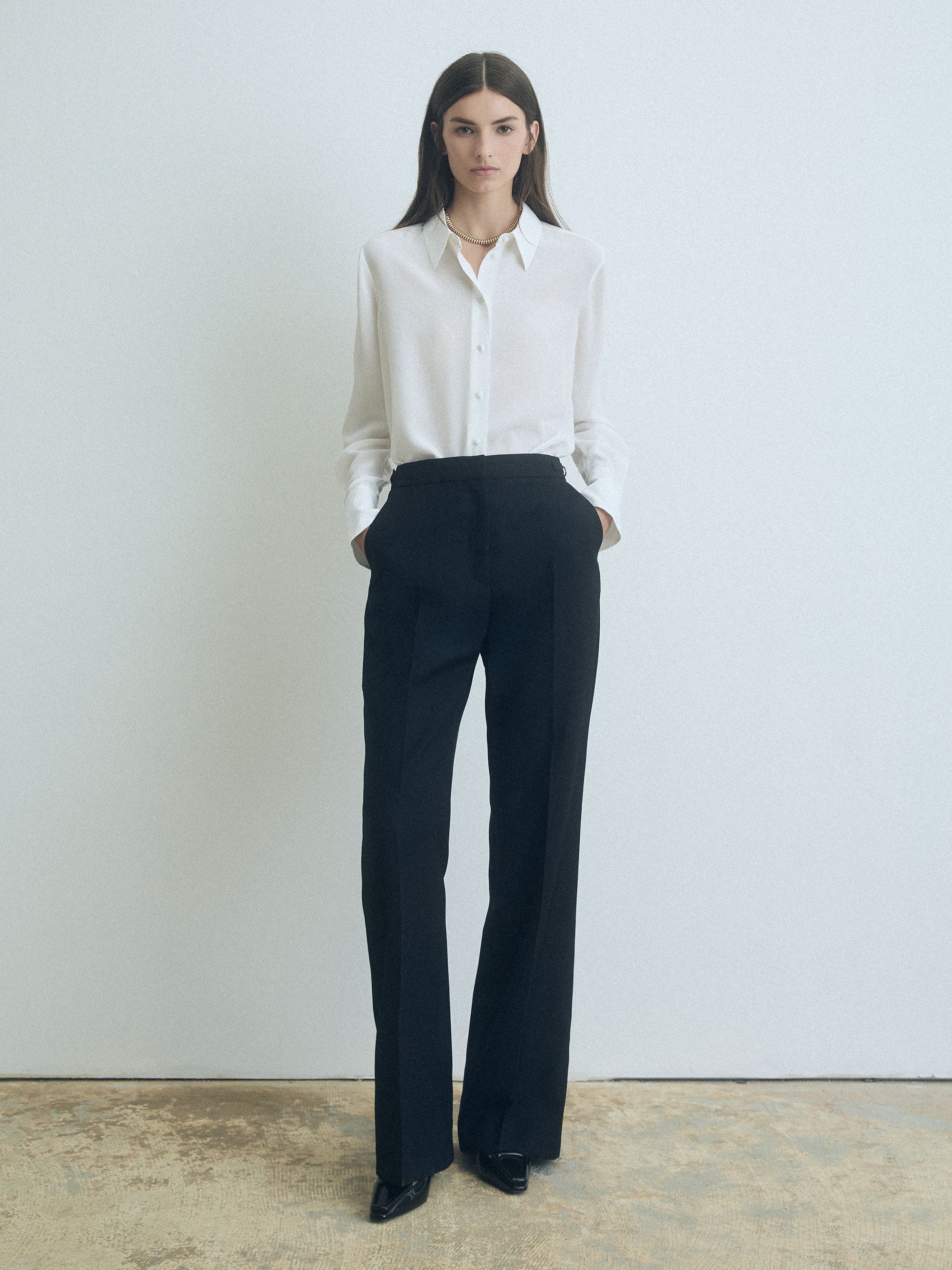 Woman wearing an off white judithj and charles blouse and black pants against a plain background