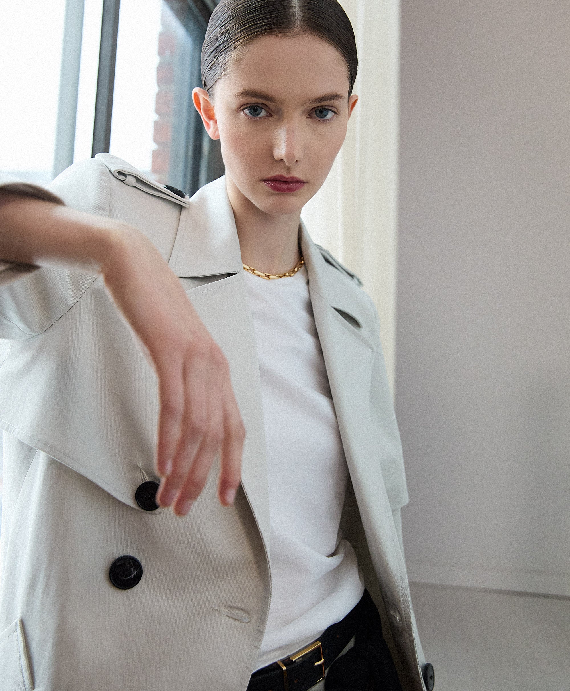 Pearl trench coat layered over a white top and dark tailored trousers, featuring double‑breasted buttons and clean structured lines in a modern indoor setting.
