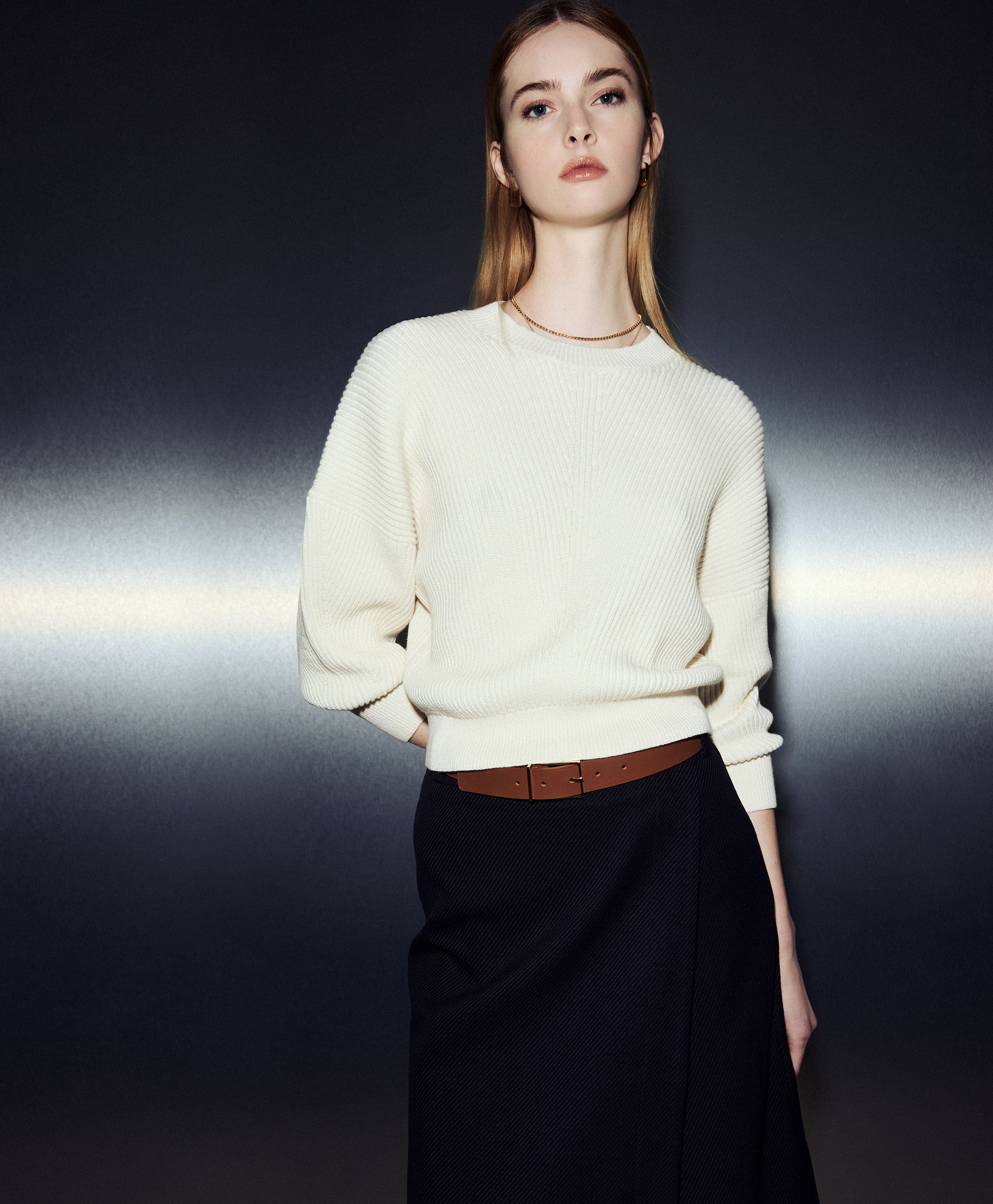 Person wearing an ivory textured knit sweater paired with a dark wrap skirt and slim belt, highlighting Judith & Charles spring knitwear in a minimal studio setting