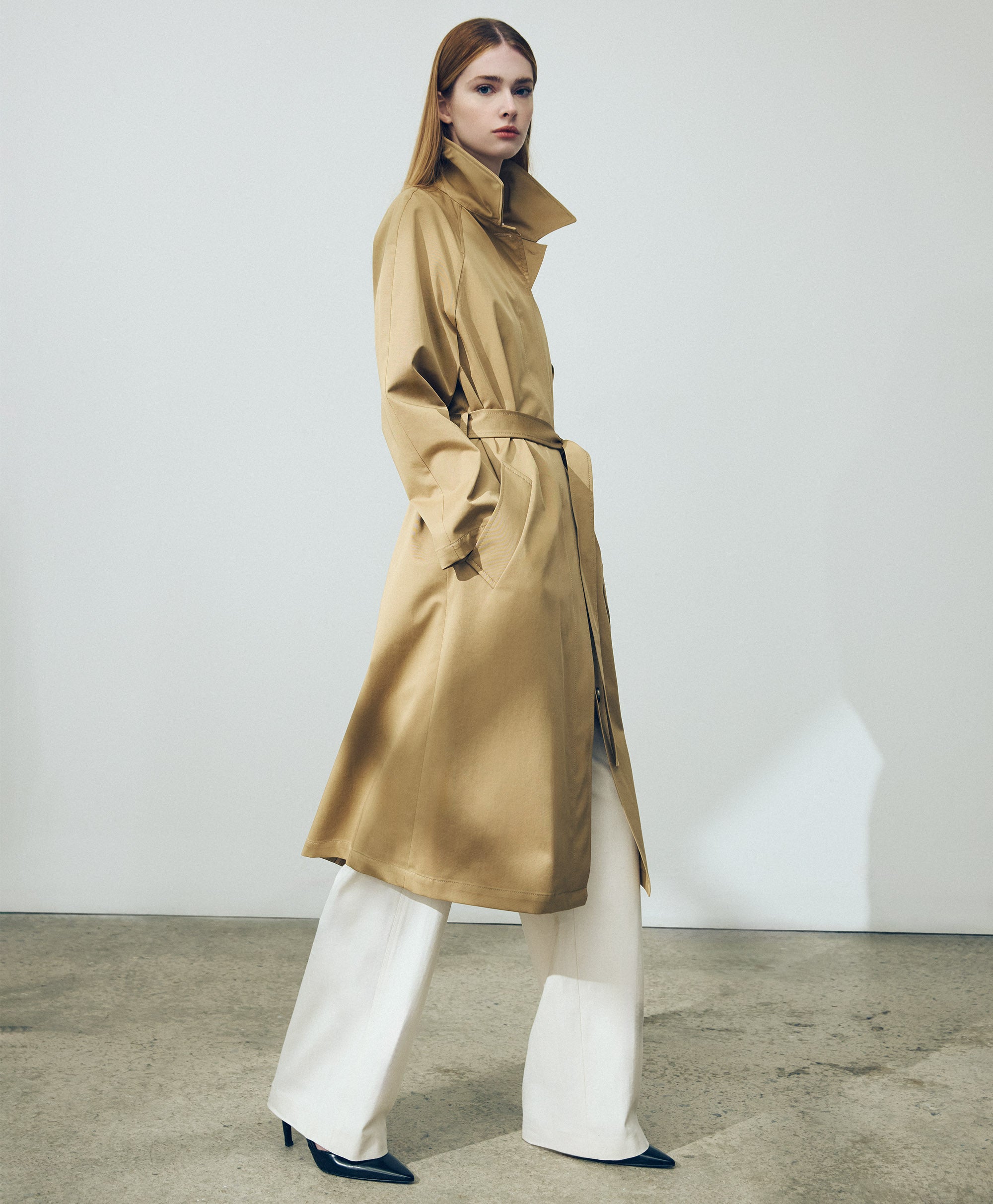 Person wearing a structured camel trench coat over wide white trousers, showcasing Judith & Charles modern tailoring in a clean studio setting.