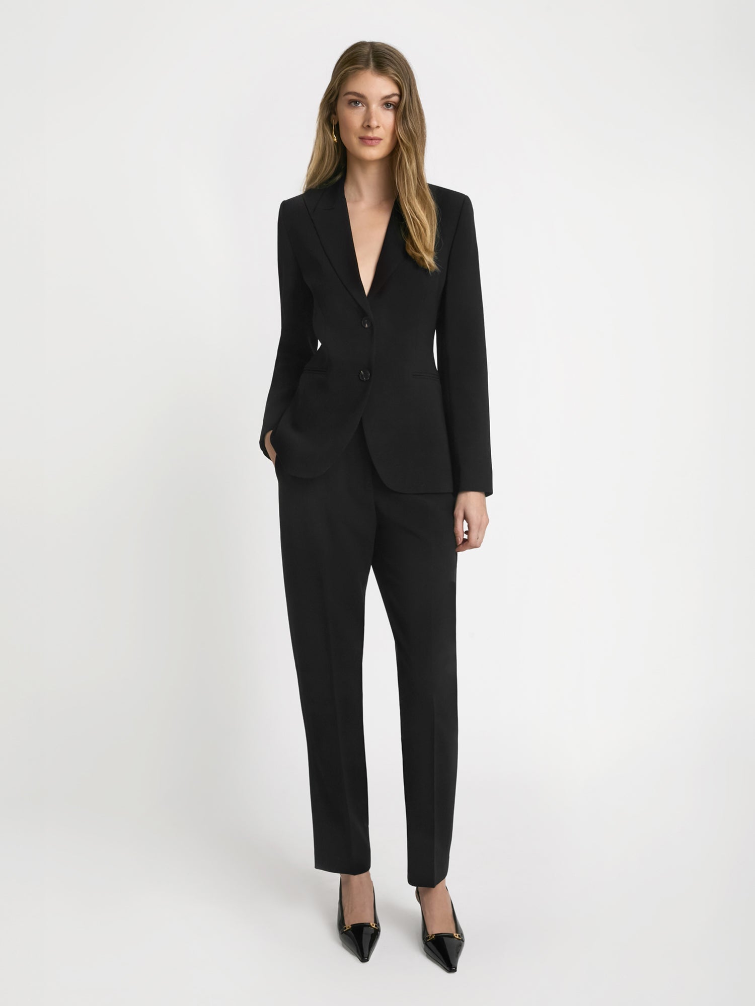 A woman standing in a studio setting, wearing a black slim pant suit with a blazer and heels.